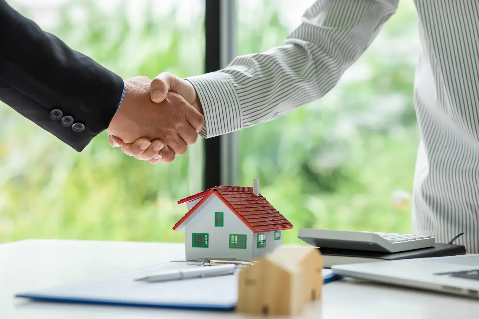 Zwei Menschen schütteln sich über einem Miniaturhaus die Hände, symbolisch für ein Immobiliengeschäft.