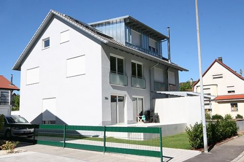 Graues zweistöckiges Haus mit grünem Zaun, geparktem Auto und blauem Himmel.