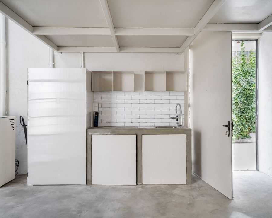 Habitación blanca vacía con un gran ventanal que da a un edificio. Suelo y techo grises.
