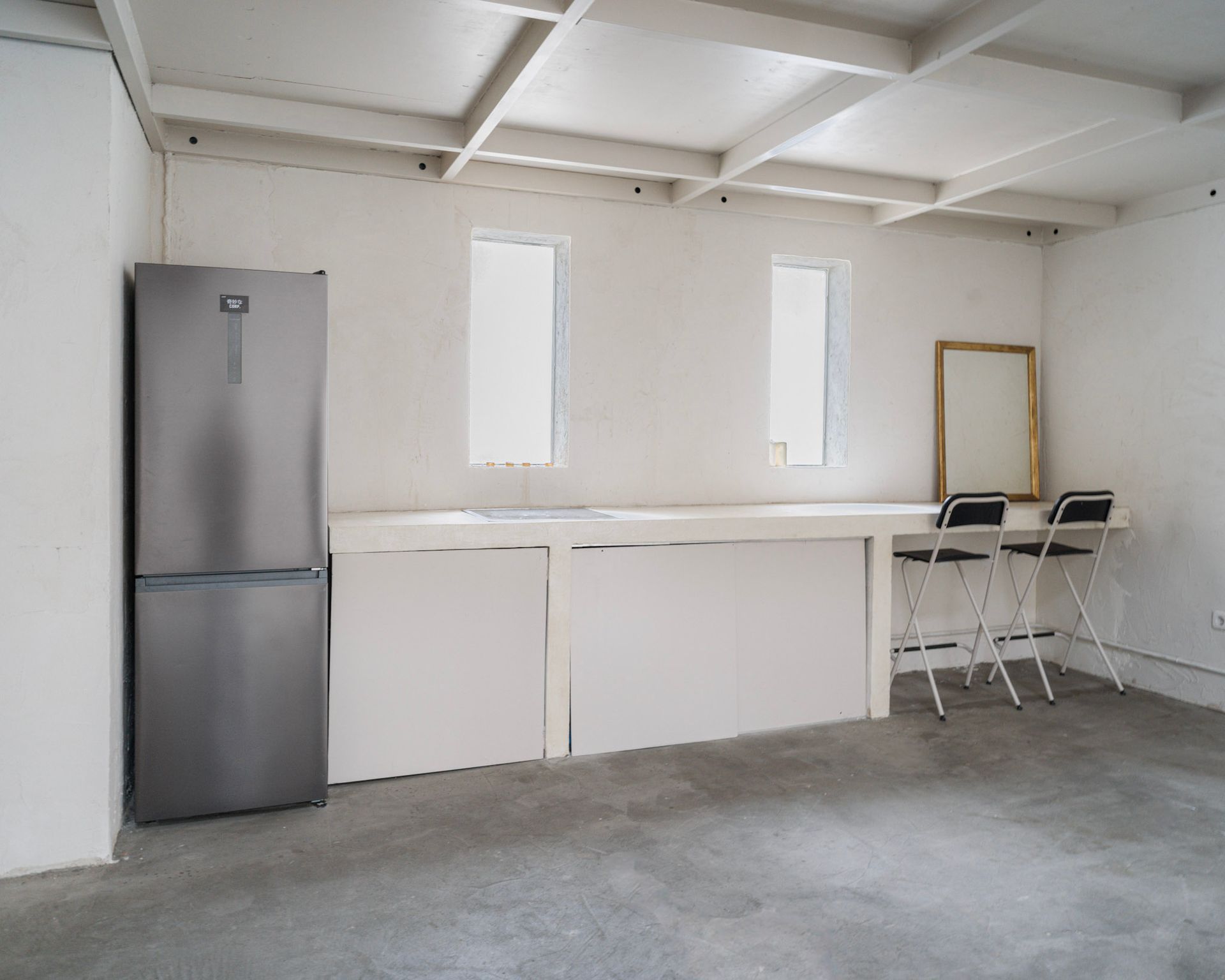 Frigorífico plateado en habitación blanca con encimera incorporada, pequeñas ventanas y dos sillas.