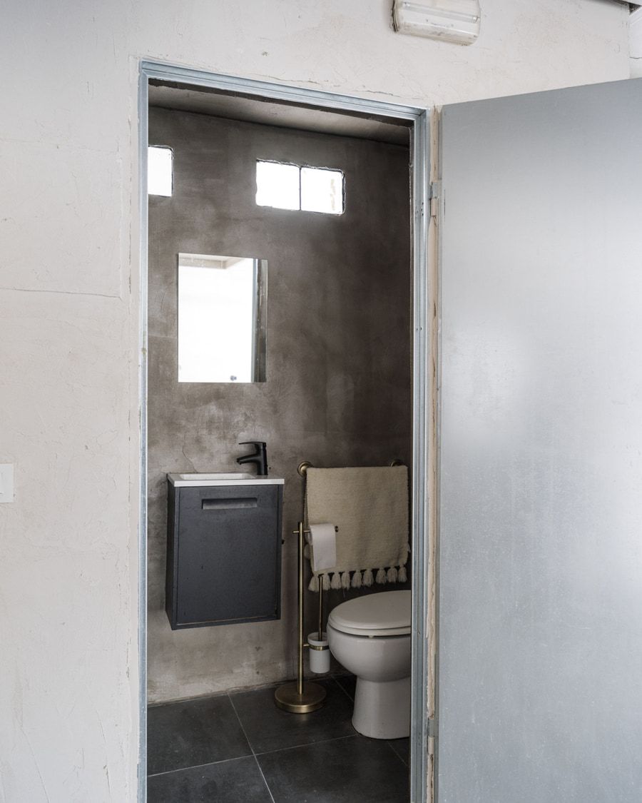 Baño estrecho con paredes grises, un pequeño tocador y un inodoro visible a través de una puerta abierta.