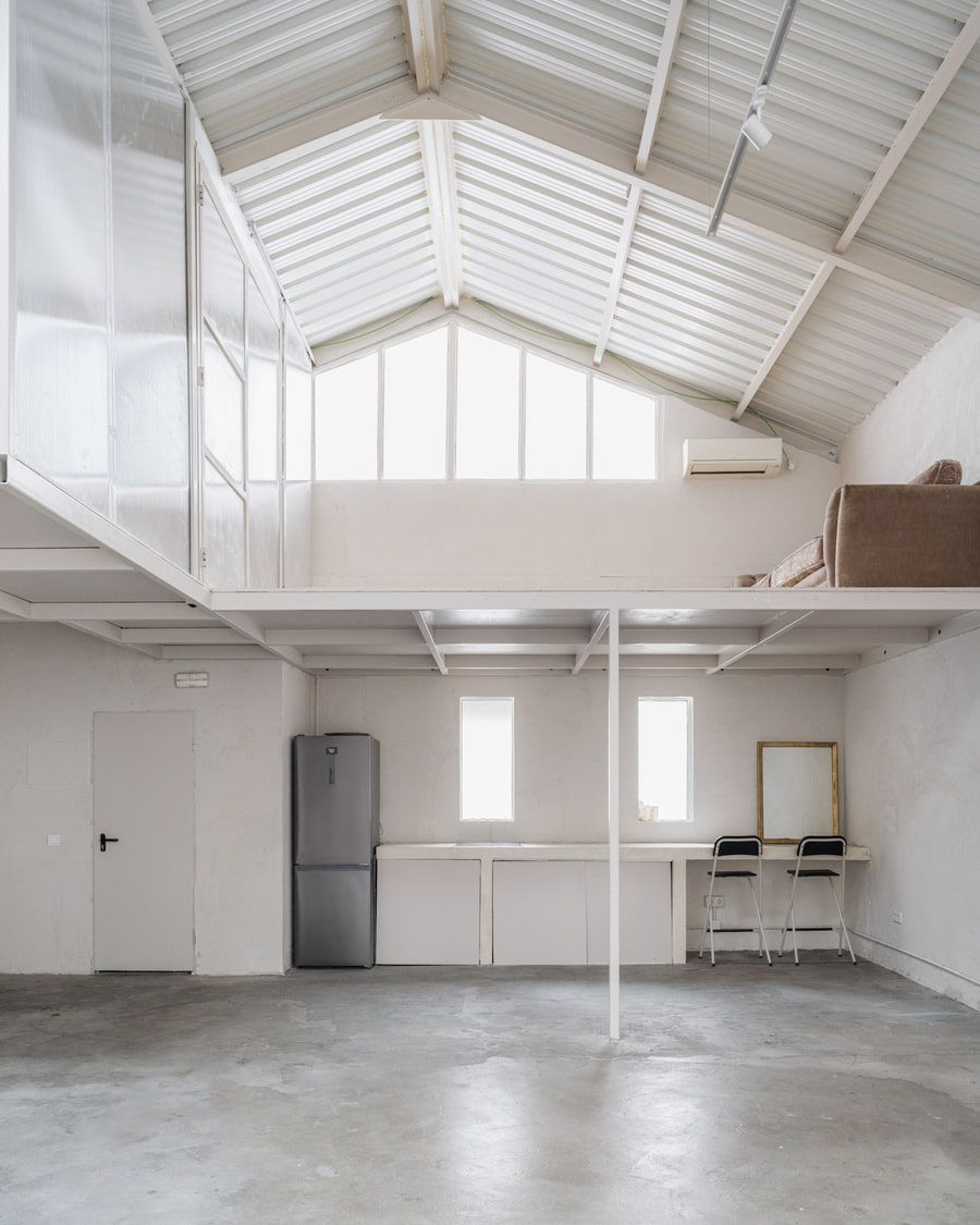 Interior de loft blanco con entrepiso, suelo de hormigón y un amplio techo inclinado. Refrigerador y escritorio.