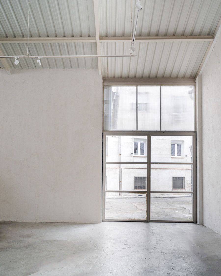 Habitación blanca vacía con un gran ventanal que da a un edificio. Suelo y techo grises.