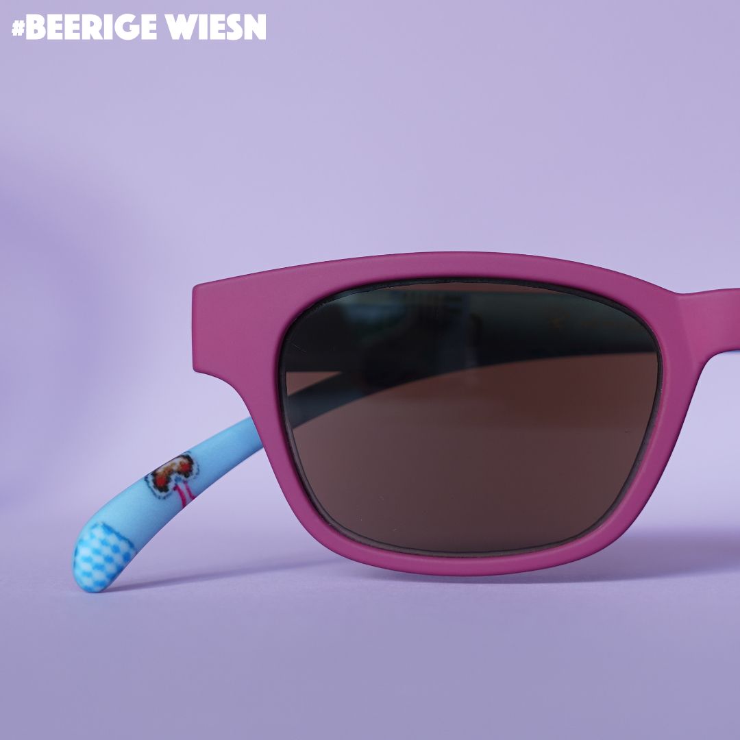 Eine rote Sonnenbrille mit blauen Bügeln im Wiesn Look auf fliederfarbenem Hintergrund.