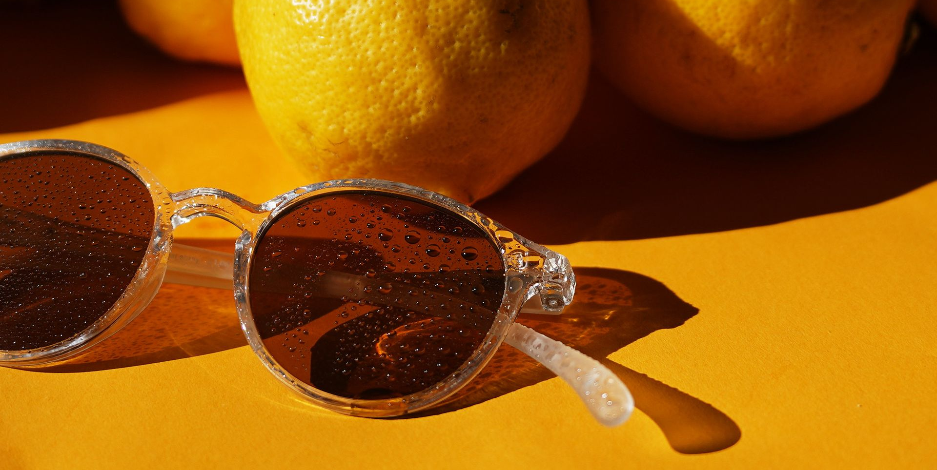 Sonnenbrille auf gelbem Untergrund mit Zitronen im Hintergrund, in Sonnenlicht getaucht.