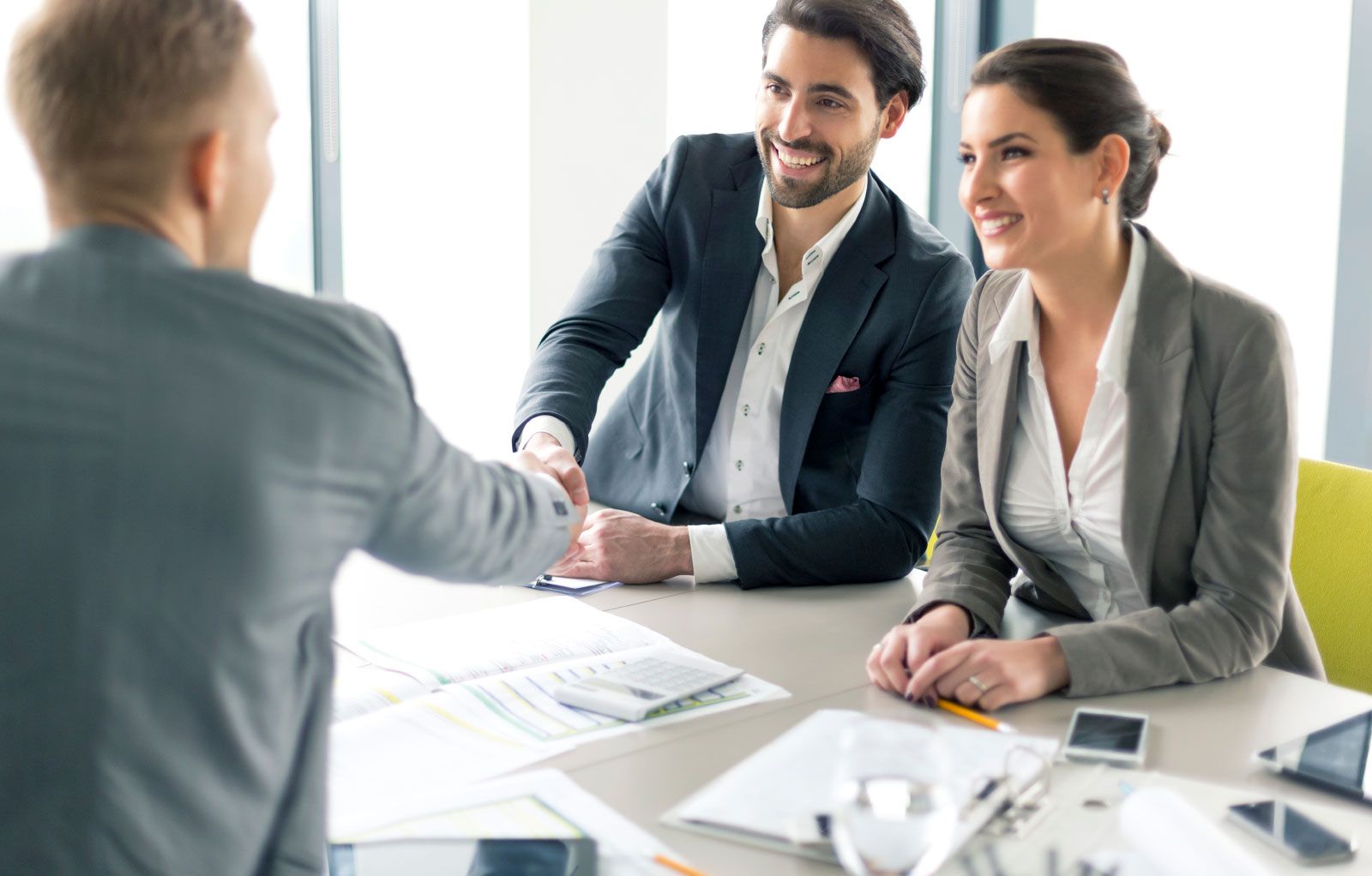 Dos personas sonriendo y dándose la mano al otro lado de un escritorio en una luminosa reunión de oficina.