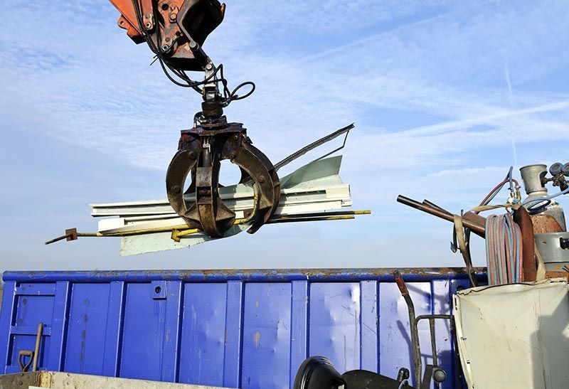 Una grúa está levantando una pila de metal hacia un contenedor de basura azul.