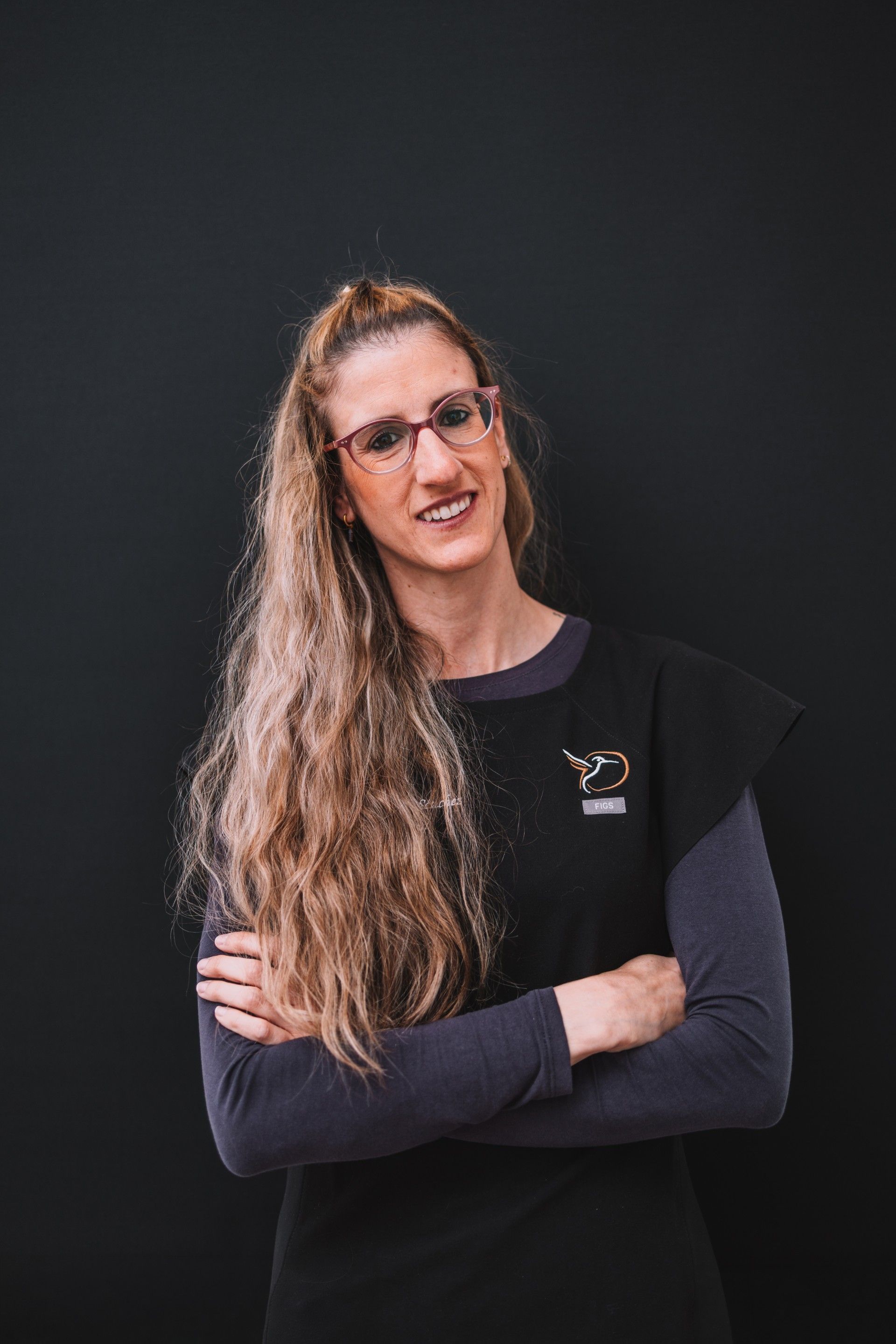Mujer con cabello largo y ondulado y gafas, de pie con los brazos cruzados sobre un fondo oscuro.