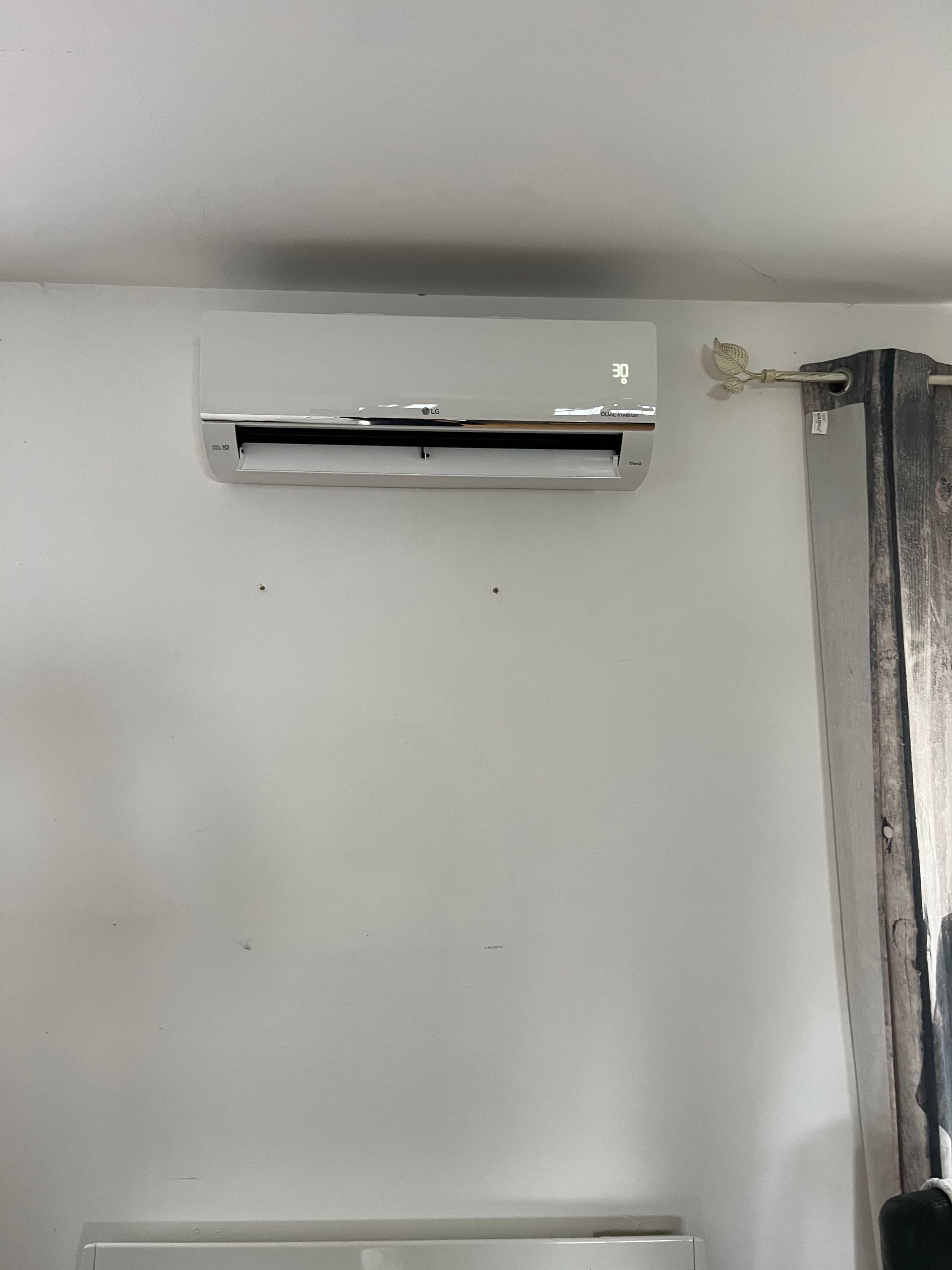 Climatiseur mural blanc au-dessus d'un mur clair uni, près d'un rideau.