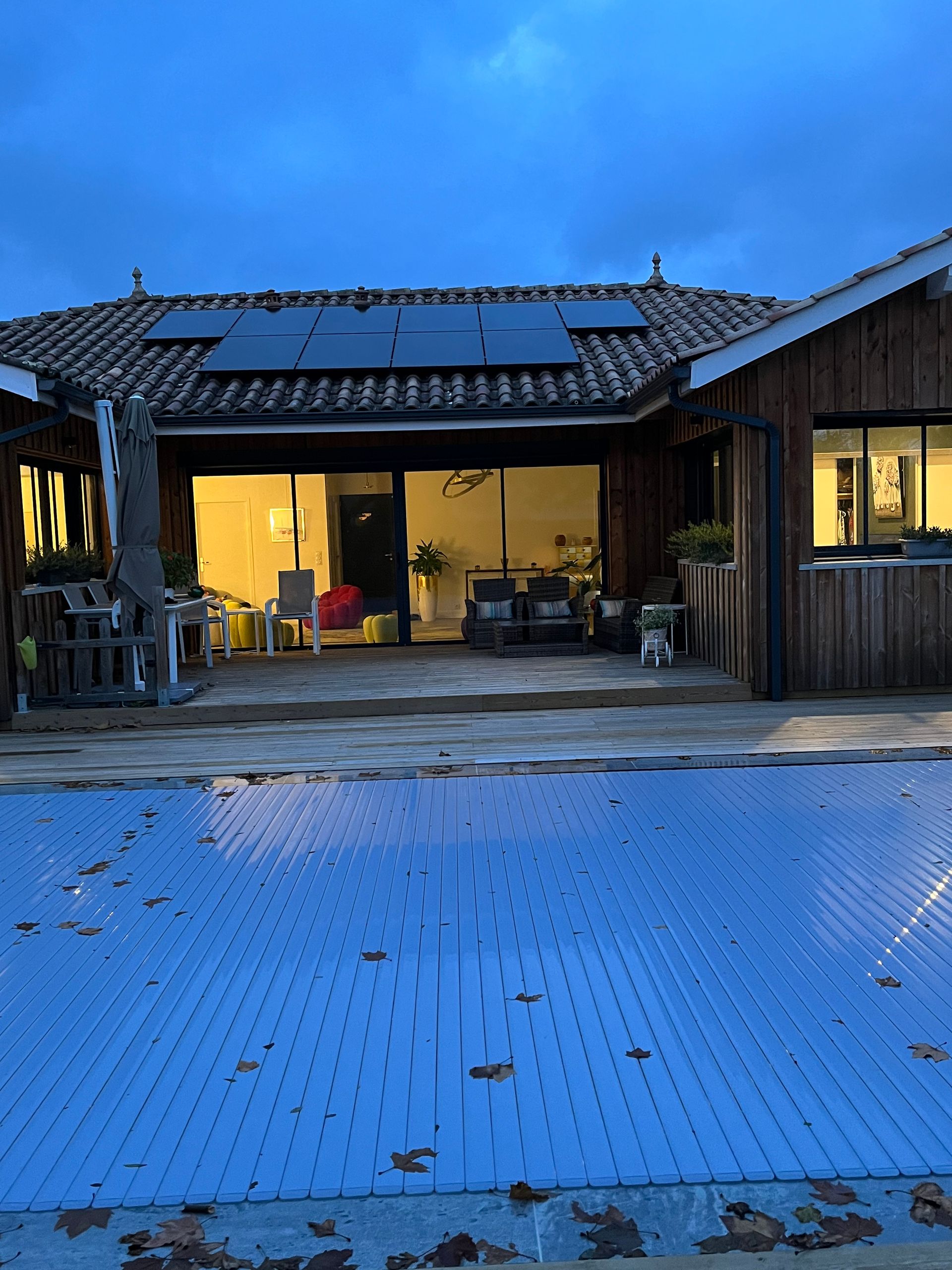 Terrasse enneigée d'un chalet au crépuscule, éclairée par des lumières chaudes à l'intérieur et un panneau solaire sur le toit