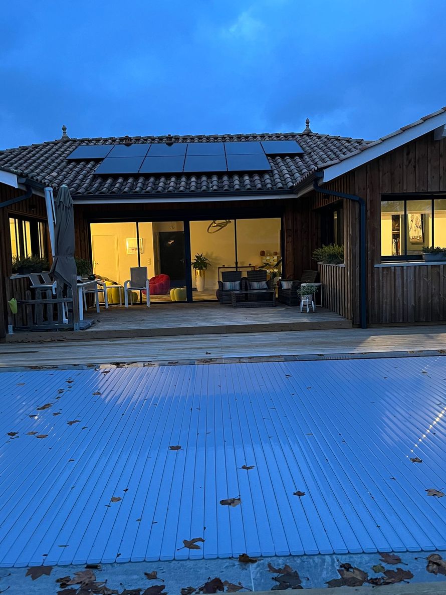 Petite maison beige avec un toit rouge, des panneaux solaires et un chemin de briques bordé de buissons taillés.