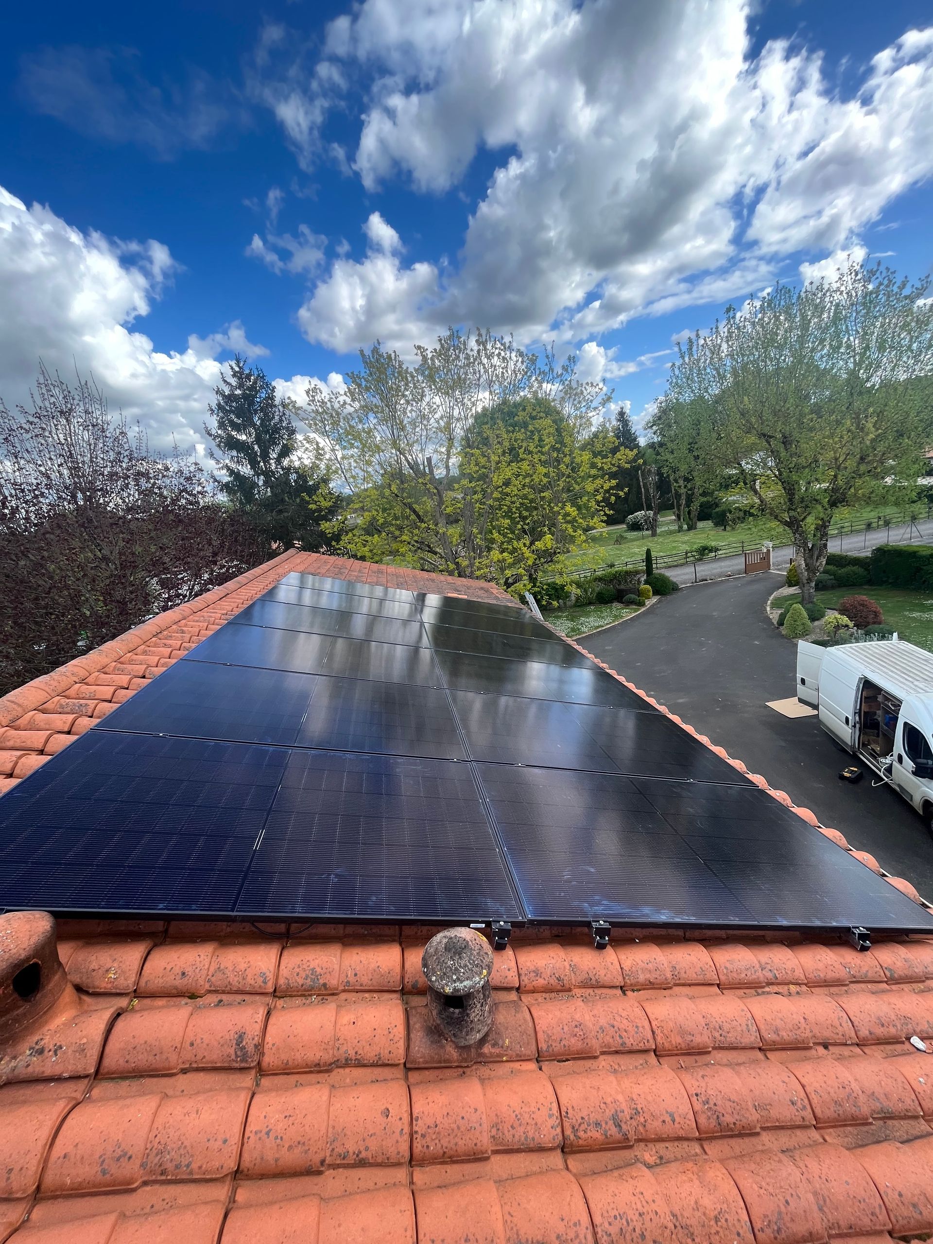Des panneaux solaires noirs sur un toit en tuiles de terre cuite sous un ciel bleu nuageux, entourés d'arbres