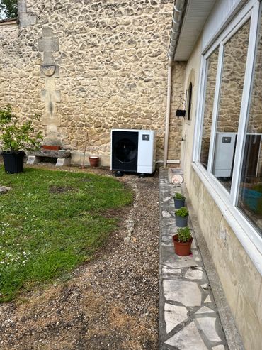 Maison en pierre avec allée de gravier, petite pelouse, plantes en pot et fenêtres le long du mur latéral.