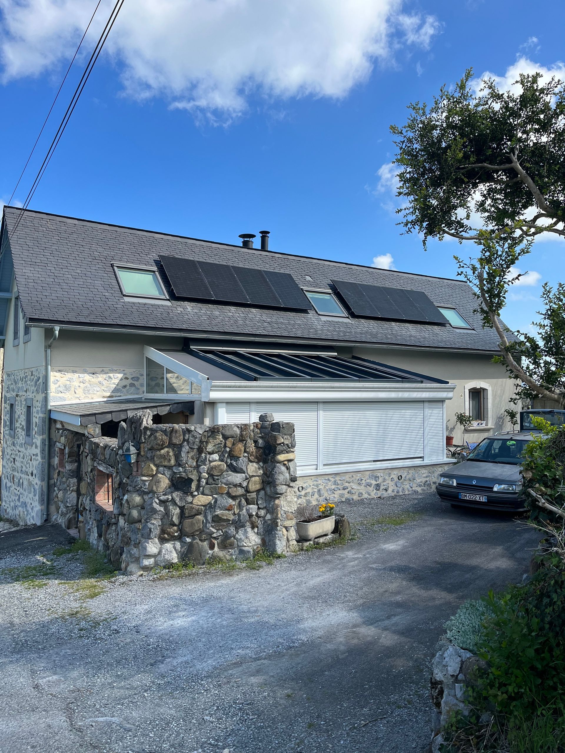 Maison en pierre avec panneaux solaires, porte de garage blanche, allée en gravier et voiture garée sous un ciel bleu