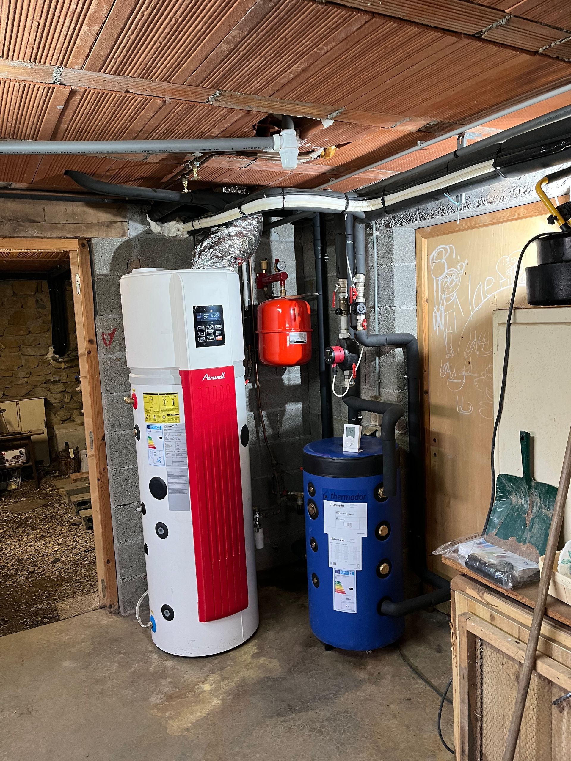 Local technique avec chauffe-eau blanc, réservoir rouge, réservoir bleu et tuyaux apparents au plafond.
