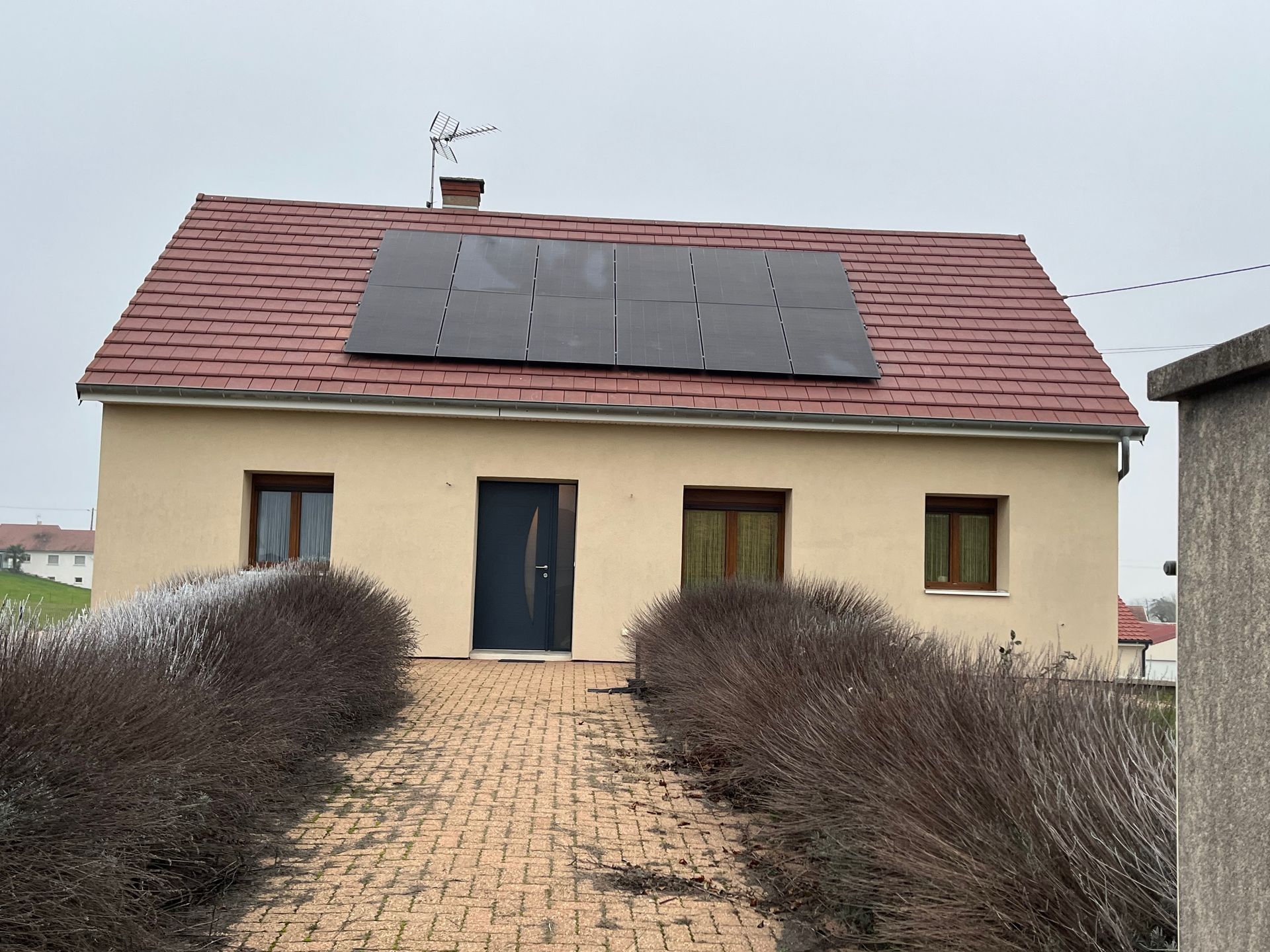 Petite maison beige avec un toit rouge, des panneaux solaires et un chemin de briques bordé de buissons taillés.