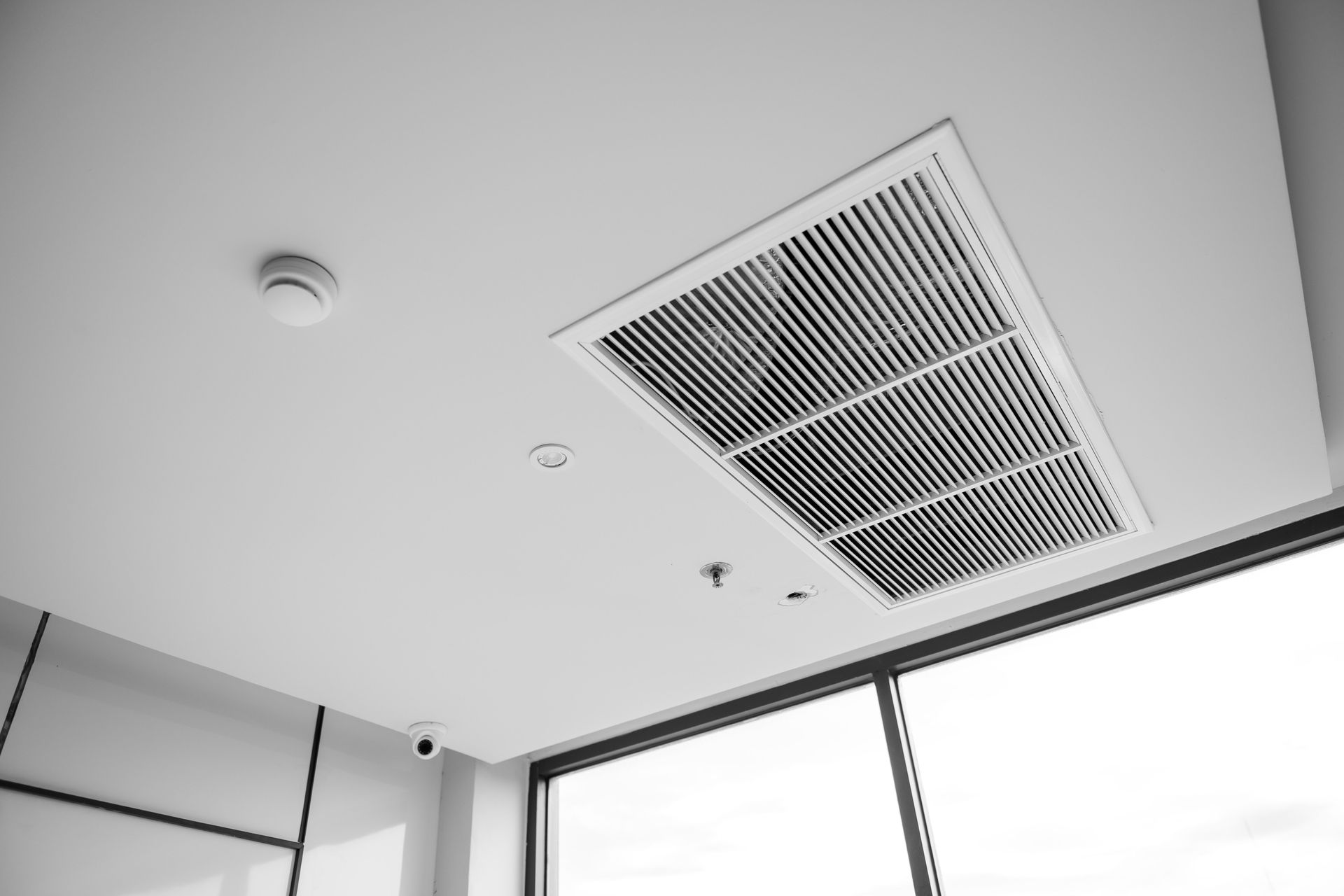 Grille d'aération au plafond et détecteur de fumée dans une pièce moderne et lumineuse avec de grandes fenêtres.