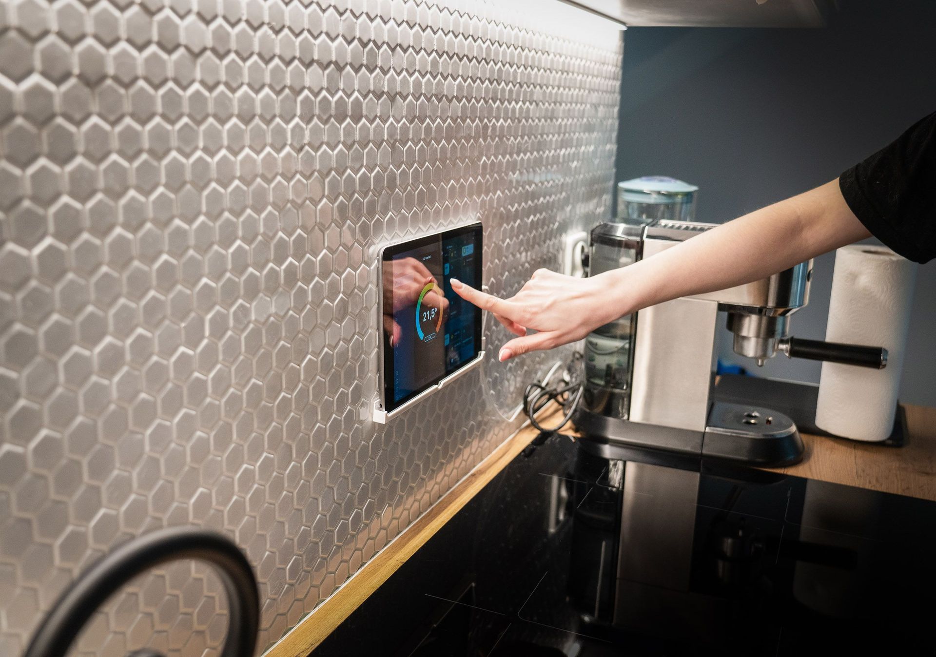 Une main se tend pour appuyer sur un écran tactile mural dans une cuisine moderne avec crédence en carreaux hexagonaux.