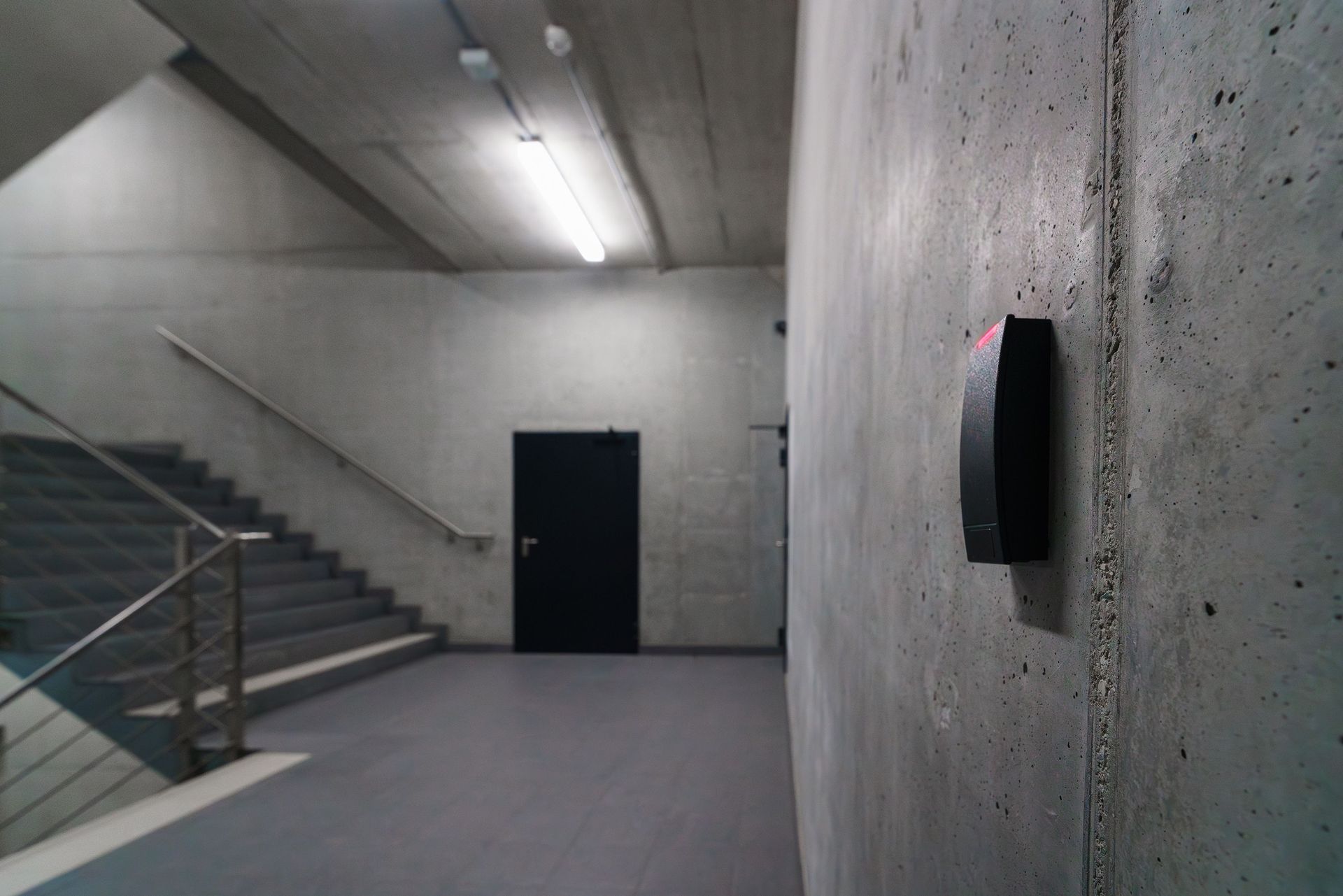 Cage d'escalier en béton avec une porte noire et un panneau d'alarme d'urgence rouge fixé au mur