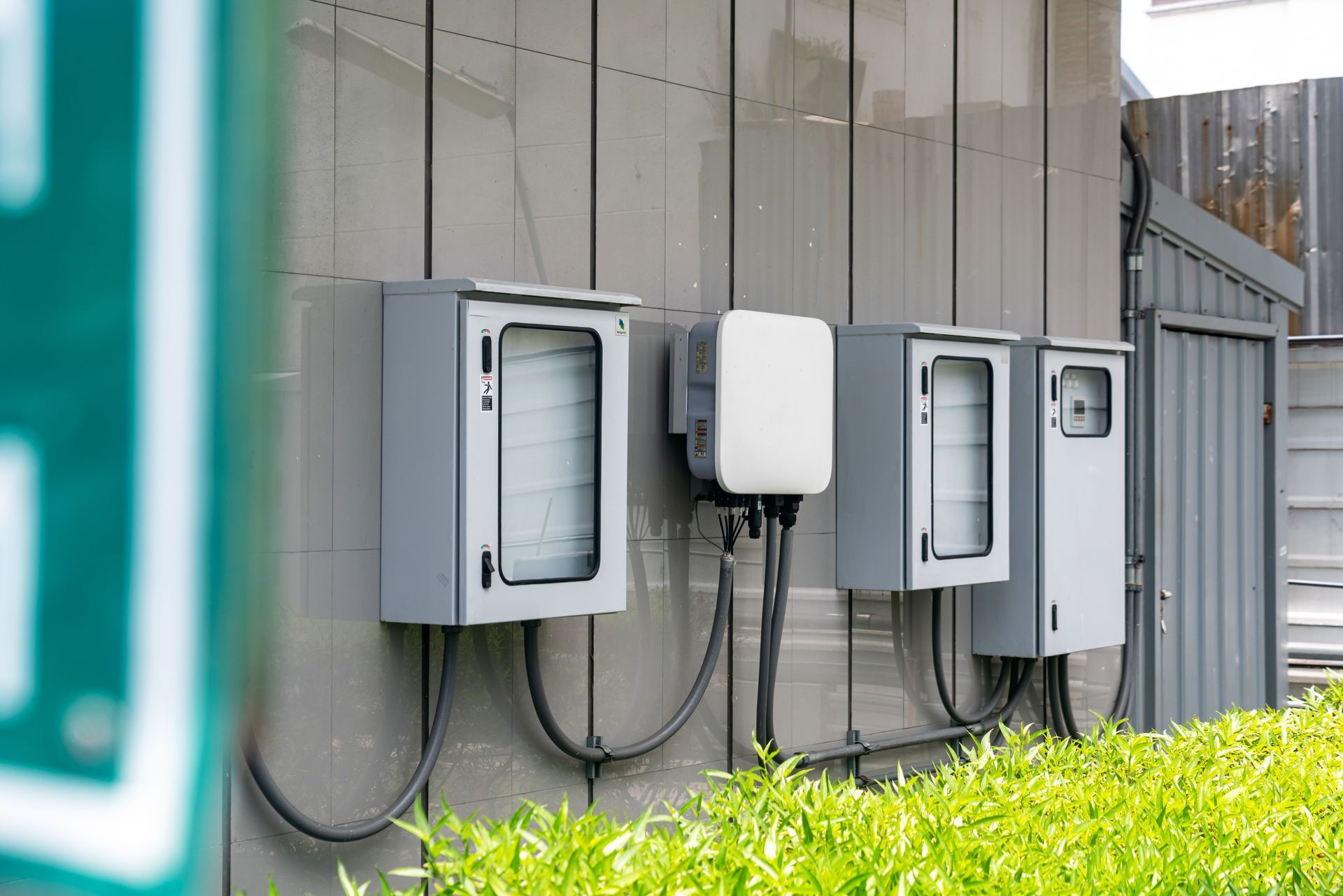 Des boîtiers électriques muraux et des équipements utilitaires jouxtent un bâtiment gris, avec des arbustes verts en contrebas.