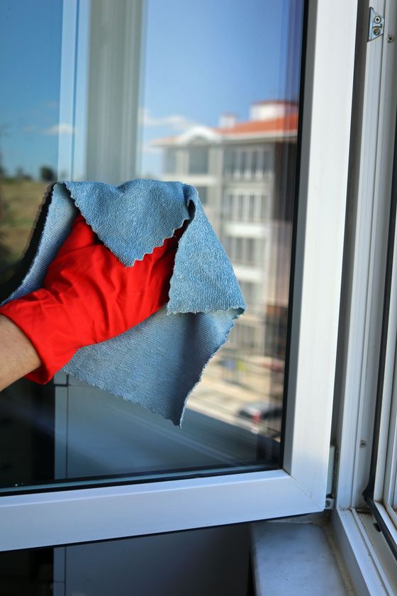 Une main gantée de rouge essuie une fenêtre avec un chiffon bleu, avec un immeuble de la ville qui se reflète à l’extérieur.