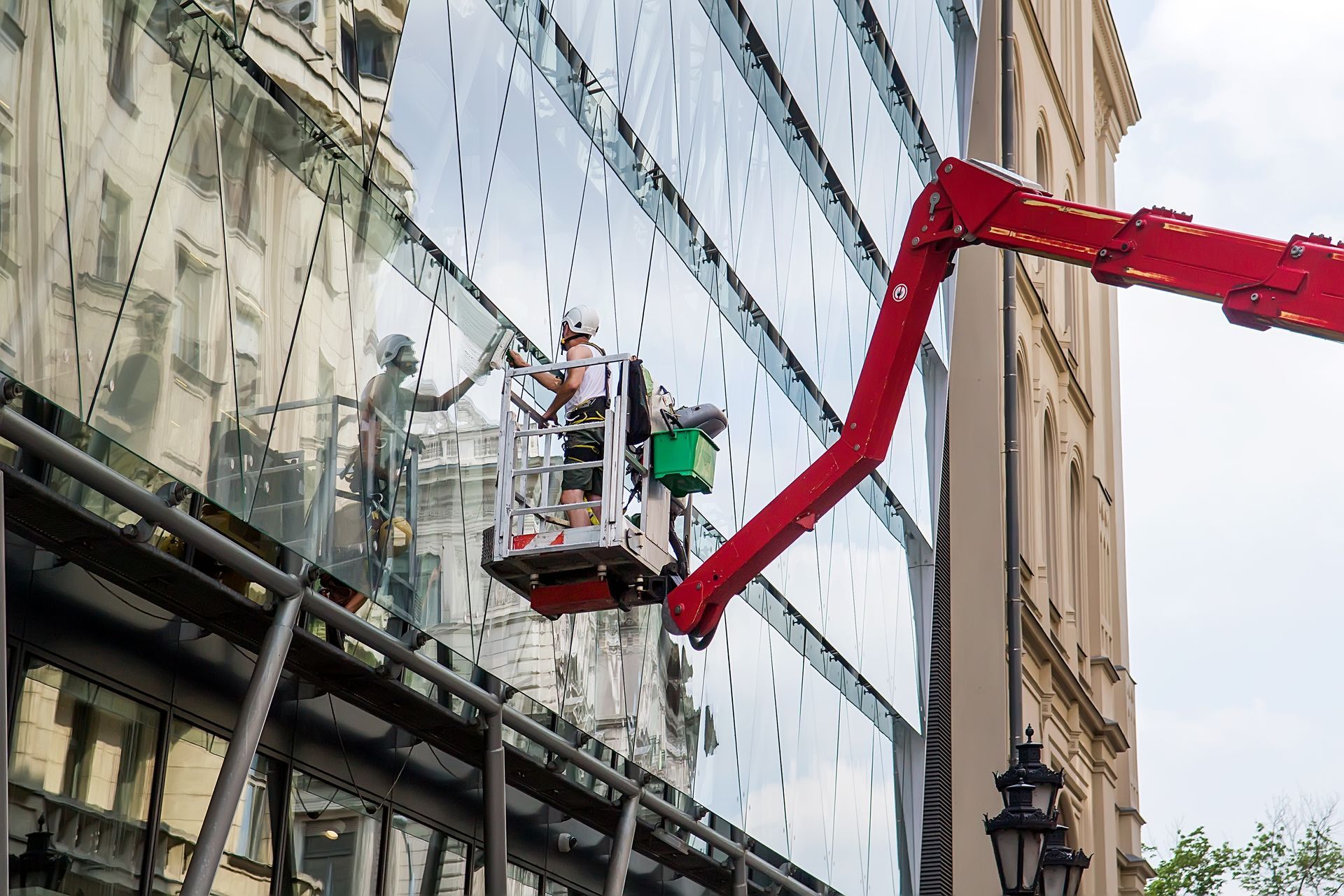 Un ouvrier dans une nacelle blanche montée sur un élévateur à bras articulé rouge nettoie la façade vitrée d’un immeuble