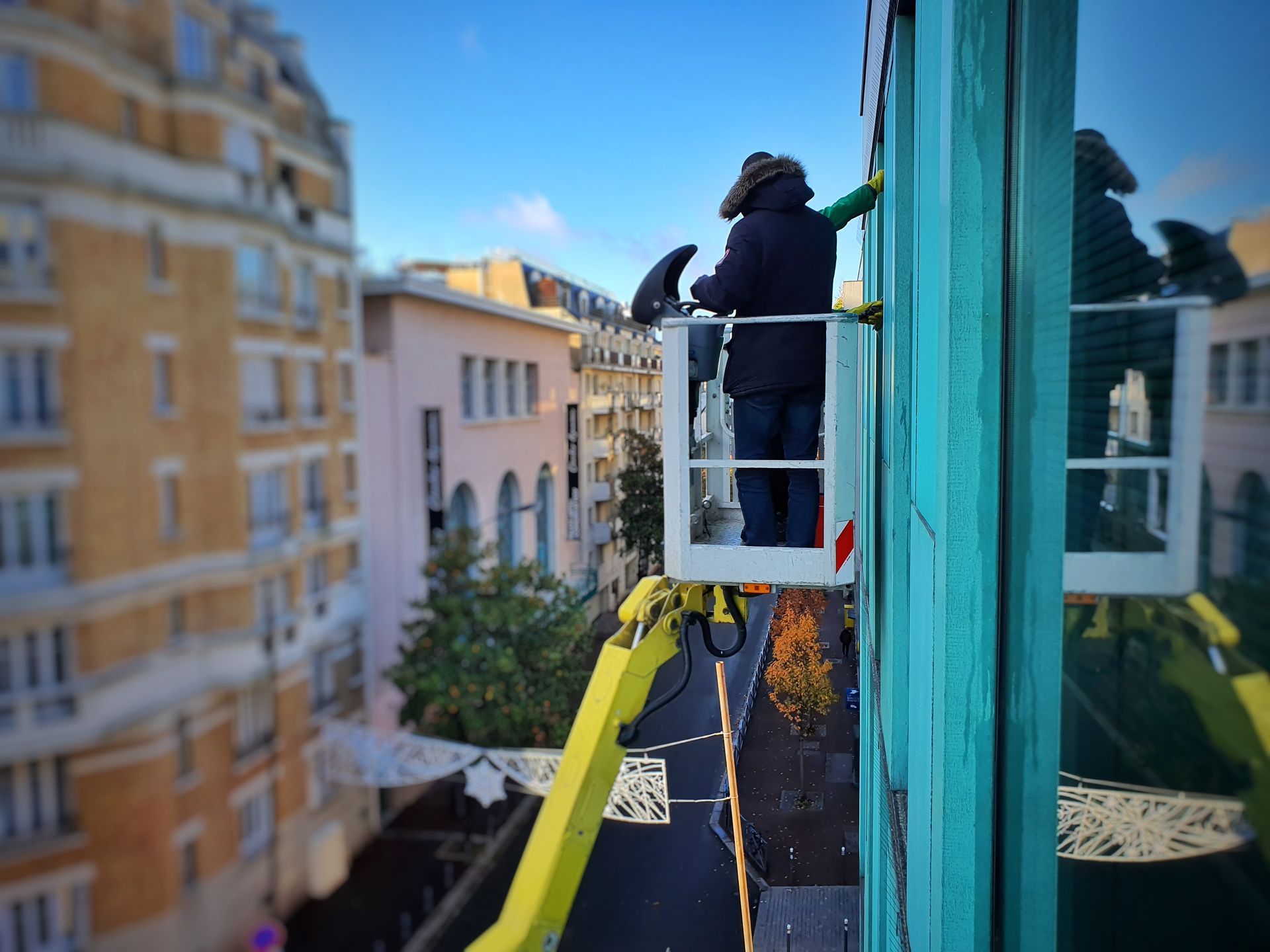 Un ouvrier sur une nacelle blanche nettoie les vitres d’un grand immeuble turquoise, avec des bâtiments en arrière-plan