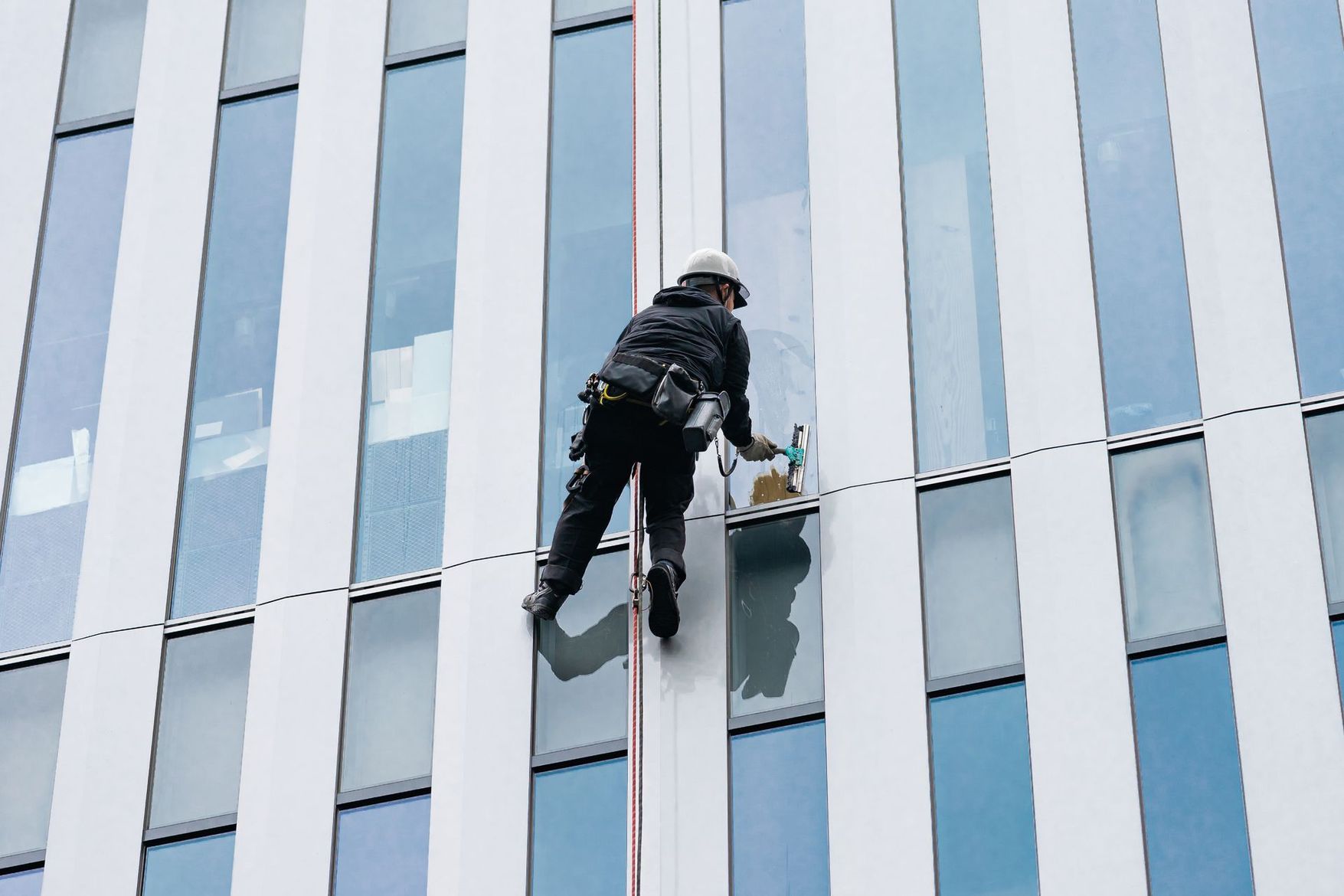Laveur de vitres suspendu à des cordes sur la façade vitrée d’un gratte-ciel, travaillant entre des panneaux teintés en bleu