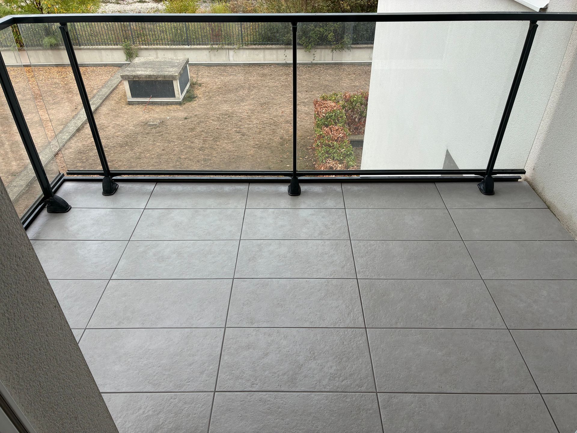 Balcon vide carrelé de gris, avec une rambarde noire donnant sur une cour et des arbres