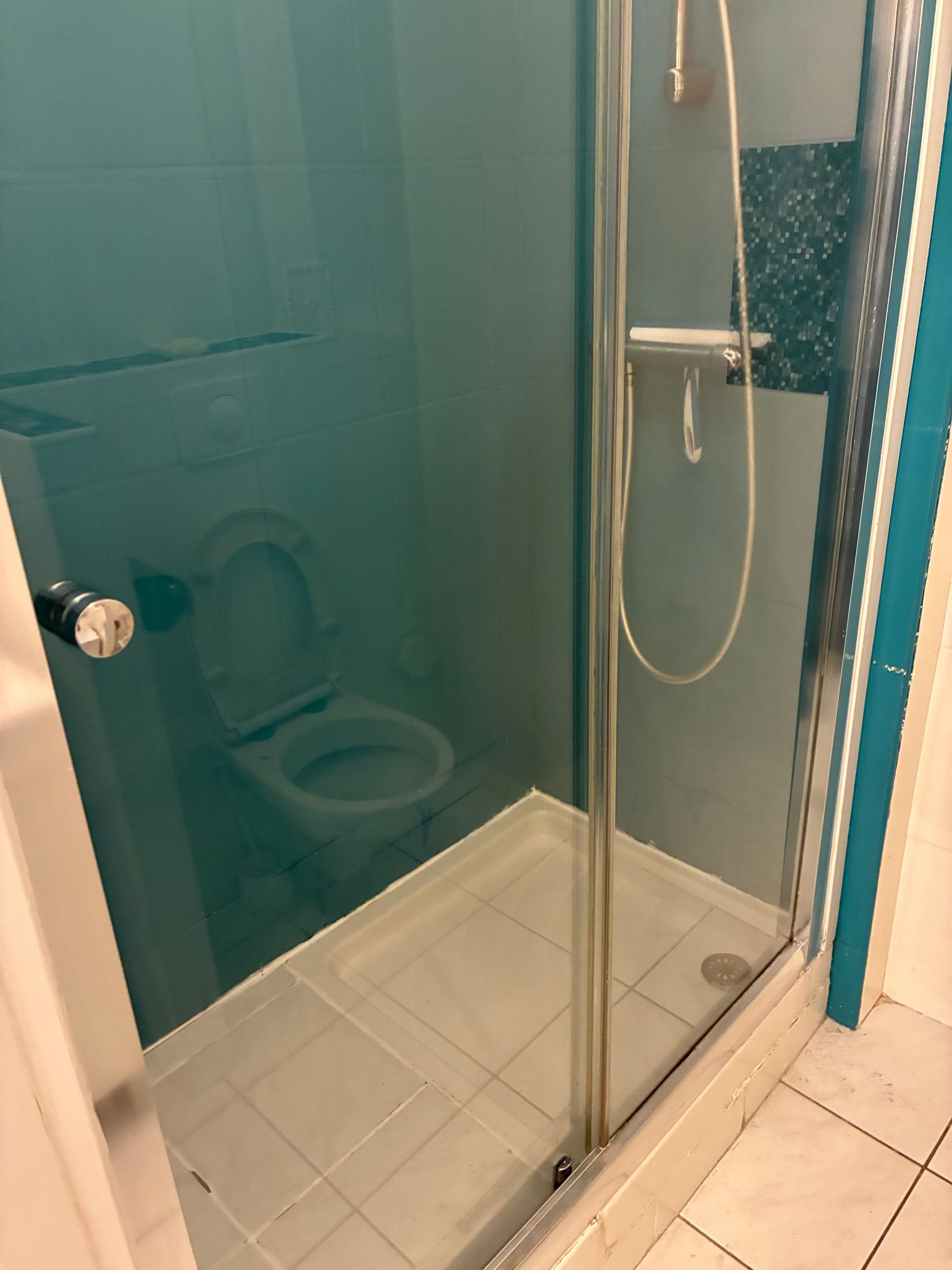 Cabine de douche en verre près d’un mur de salle de bains bleu sarcelle, avec les toilettes visibles derrière la paroi.