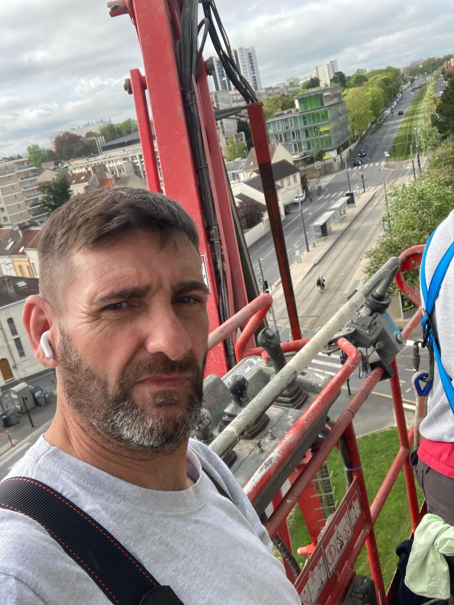 Un homme sur une nacelle de grue aux garde-corps rouges, surplombant une rue de la ville par temps nuageux