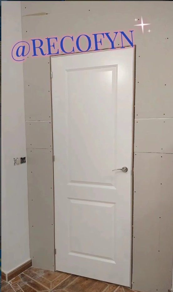 Puerta interior blanca en una pared parcialmente terminada con piso de baldosas y una manija plateada.