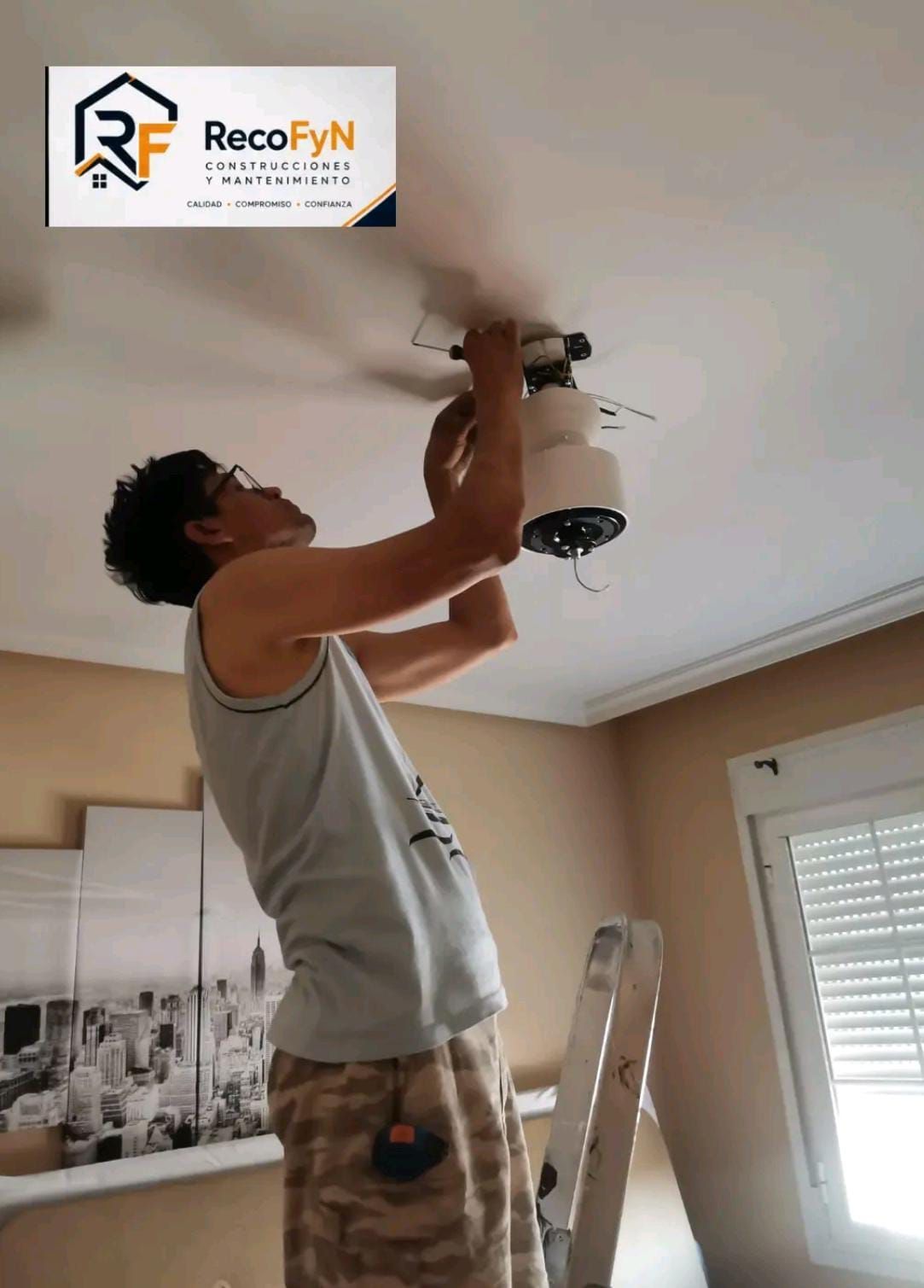 Un hombre utiliza un taladro para instalar una lámpara de techo en una habitación con techo blanco y persianas en las ventanas.