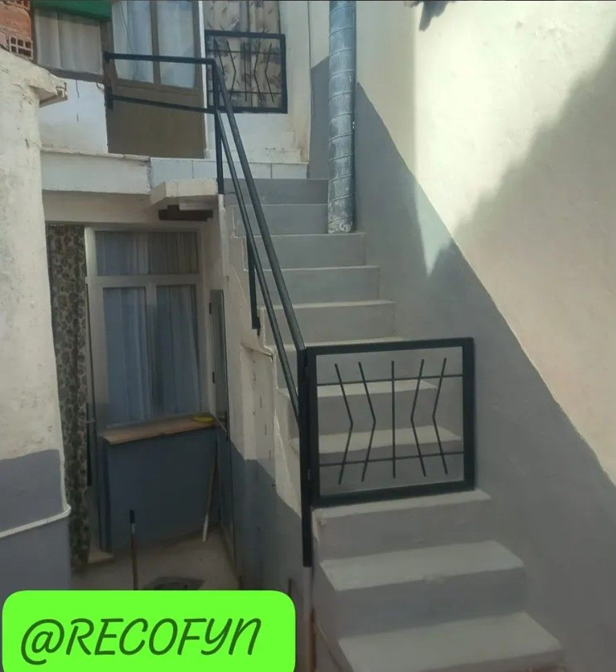 Estrecha escalera exterior entre edificios con barandilla negra y escalones grises, bañada por la luz del sol.