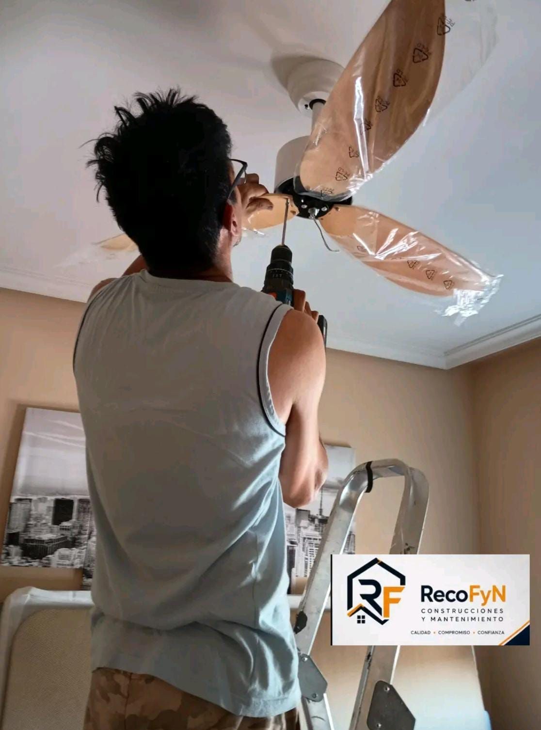 Un trabajador subido a una escalera instala un ventilador de techo en una habitación, con las aspas del ventilador envueltas en plástico protector.