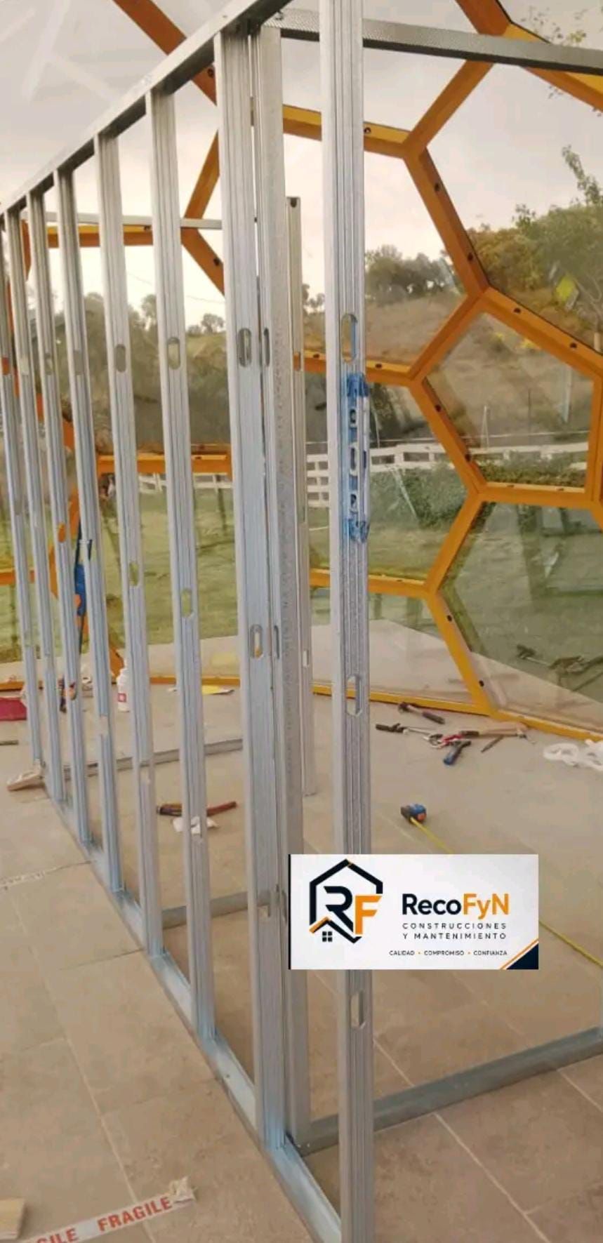 Estructura metálica para un cerramiento de vidrio en un patio de concreto, con una ventana y un edificio de color marrón anaranjado al fondo.