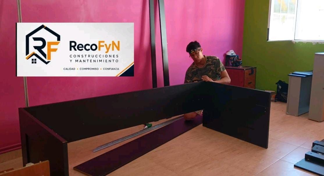 Una persona está montando un escritorio negro en una habitación con una pancarta de RecoFyN en la pared.