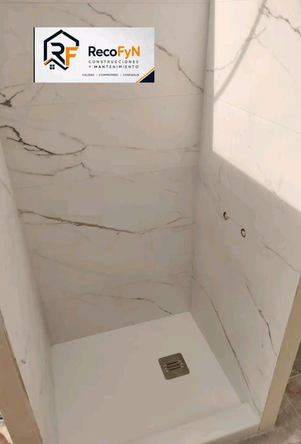 Cabina de ducha vacía con azulejos, paredes con aspecto de mármol, base blanca y desagüe en el suelo.