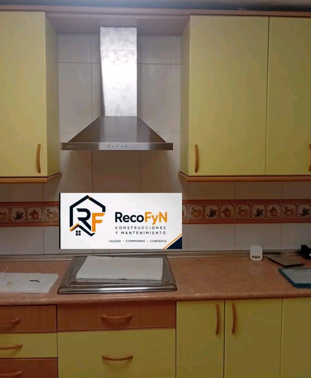 Cocina amarilla con campana extractora de acero inoxidable, encimera y puertas de armarios de color beige, además de un letrero de RecoFYN cerca del fregadero.