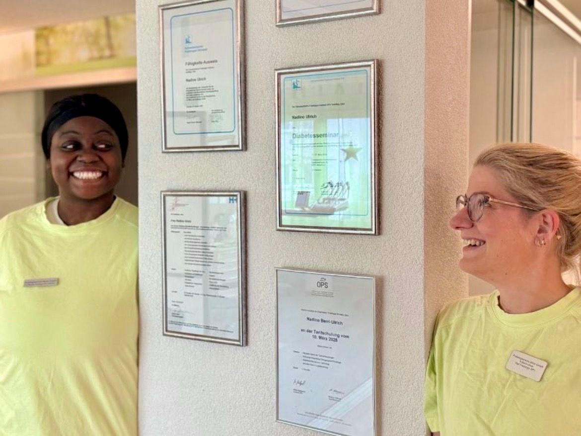 Zwei Personen in hellgrünen Hemden lächeln vor gerahmten Urkunden an einer Wand.