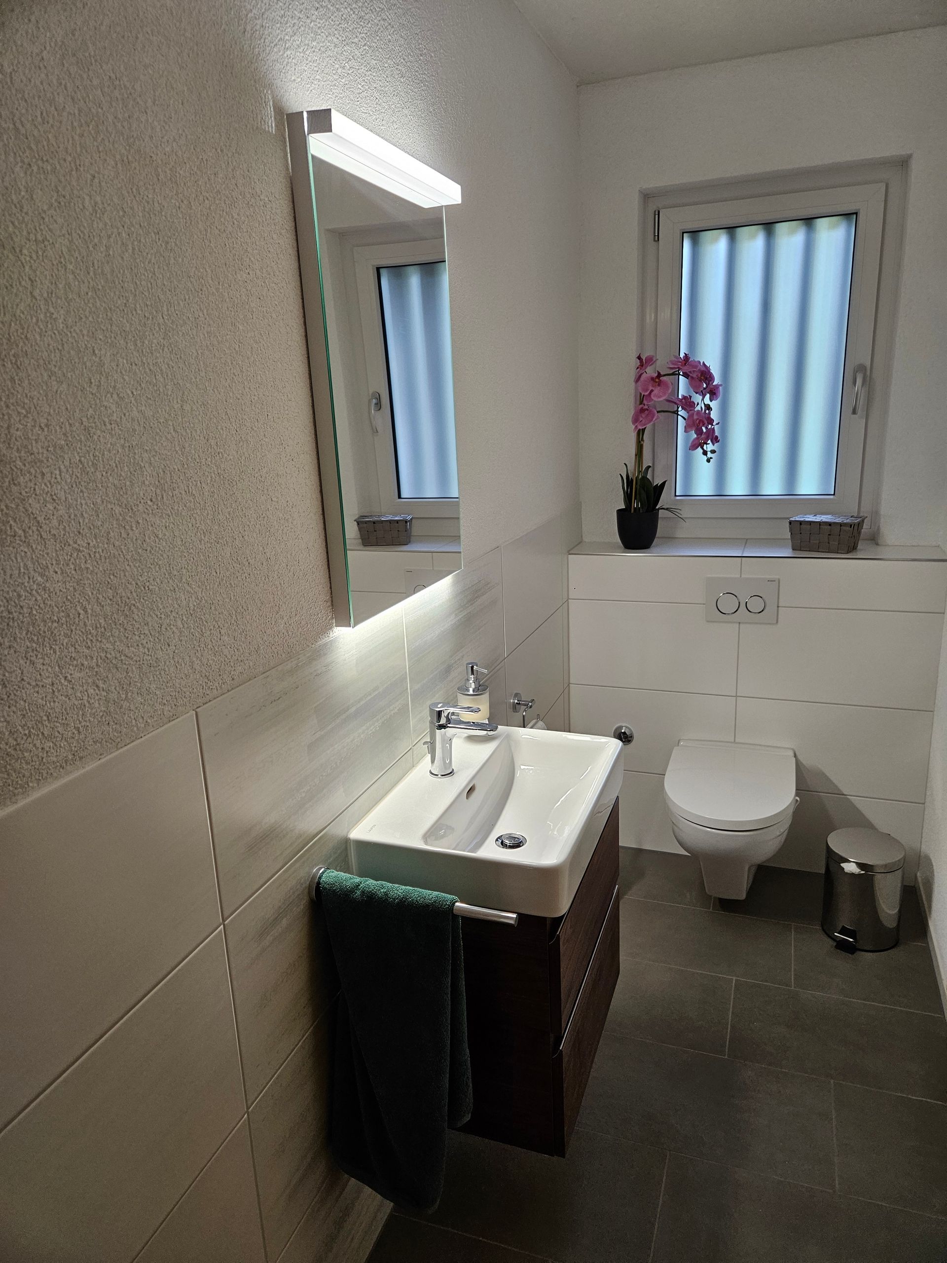 Badezimmer mit Duschkabine aus Glas, gefliesten Wänden und Boden, Fenster und Waschtisch.