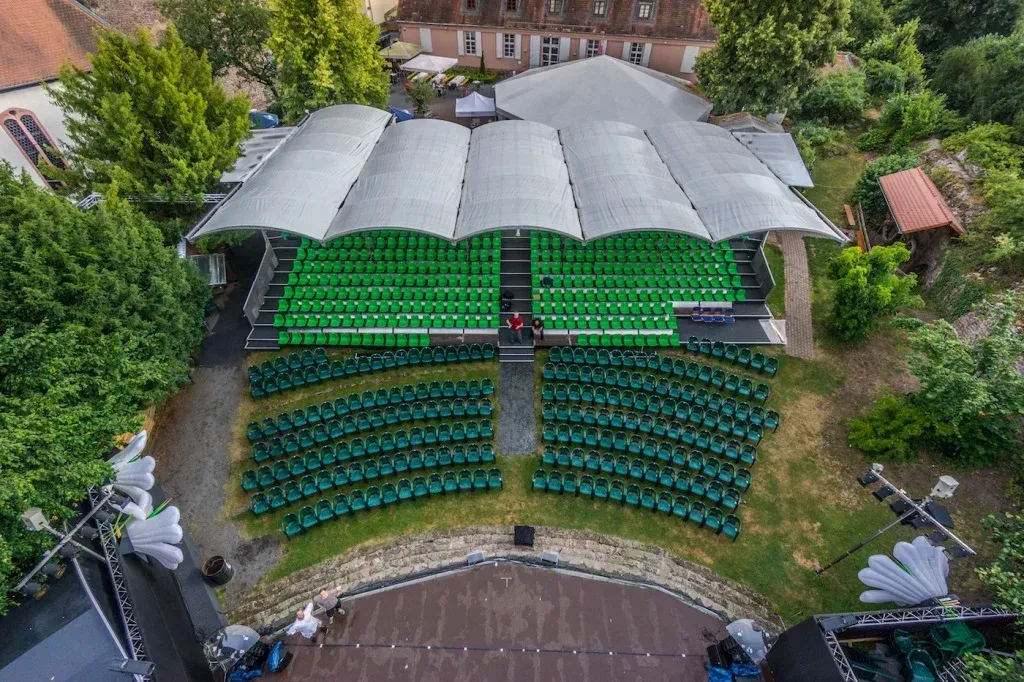 Draufsicht auf grüne Sitzplätze in einem Freiluft-Amphitheater, überdacht von einer weißen Markise und umgeben von Bäumen.