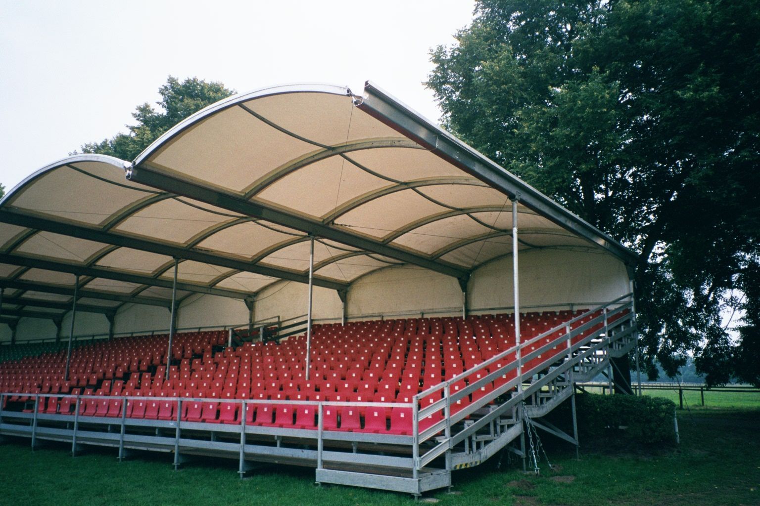 Rote Stadionsitze unter einem weißen Bogendach, in einer Außenumgebung mit Bäumen.