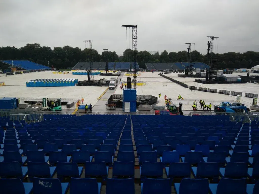 Blaue Stadionsitze blicken auf eine im Aufbau befindliche Bühne, unter einem bewölkten Himmel sind Arbeiter und Ausrüstung zu sehen.