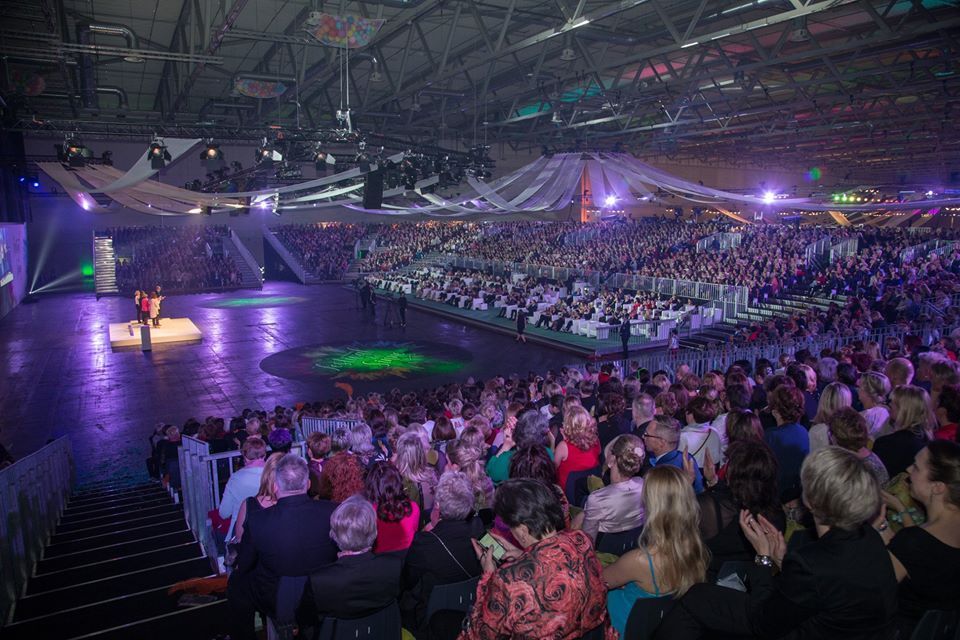 Das Publikum beobachtet eine Aufführung in einer großen, beleuchteten Arena. Links auf der Bühne ist ein Künstler beleuchtet.