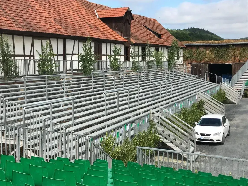 Tribüne im Freien vor einem traditionellen Fachwerkhaus. In der Nähe parkt ein weißes Auto.
