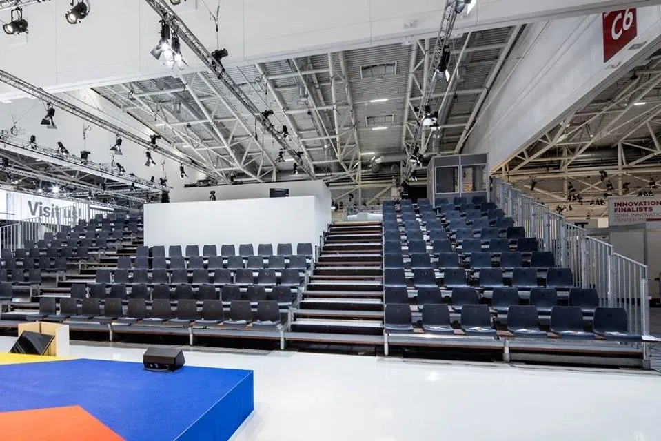 Tribüne in einer Halle. Leere, graue Sitze. Bühnenbild mit einer blauen Matte davor.