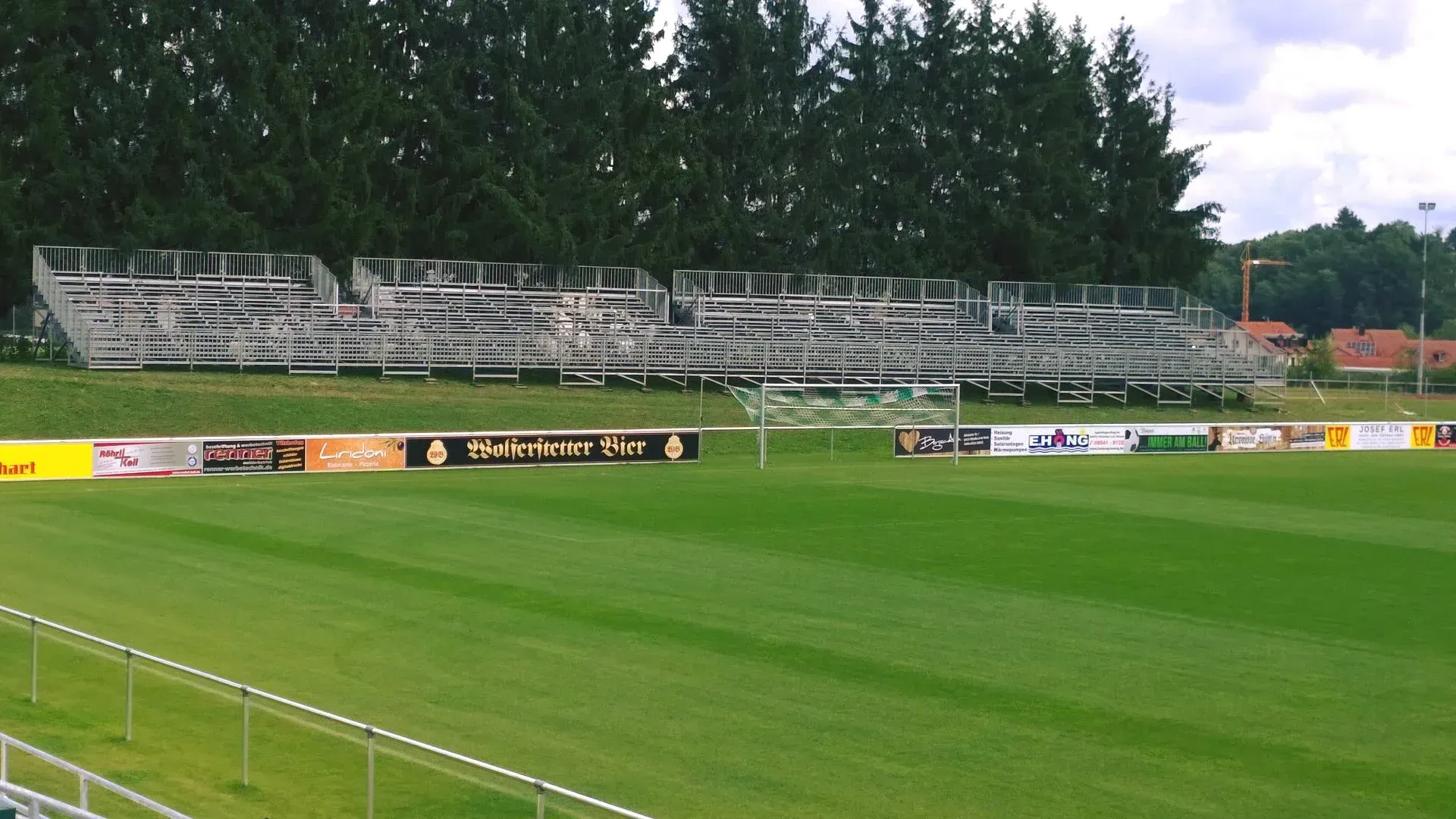 Leerer Fußballplatz mit Metalltribünen, Werbebannern und einer Baumreihe.