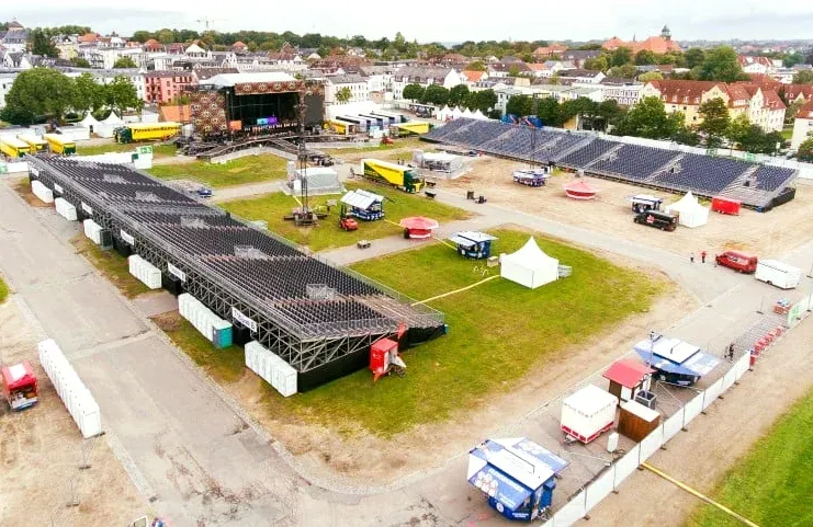 Aufbau eines Musikfestivals im Freien mit Bühne, Sitzgelegenheiten, Verkaufszelten und Ausrüstung auf einem Stadtplatz.