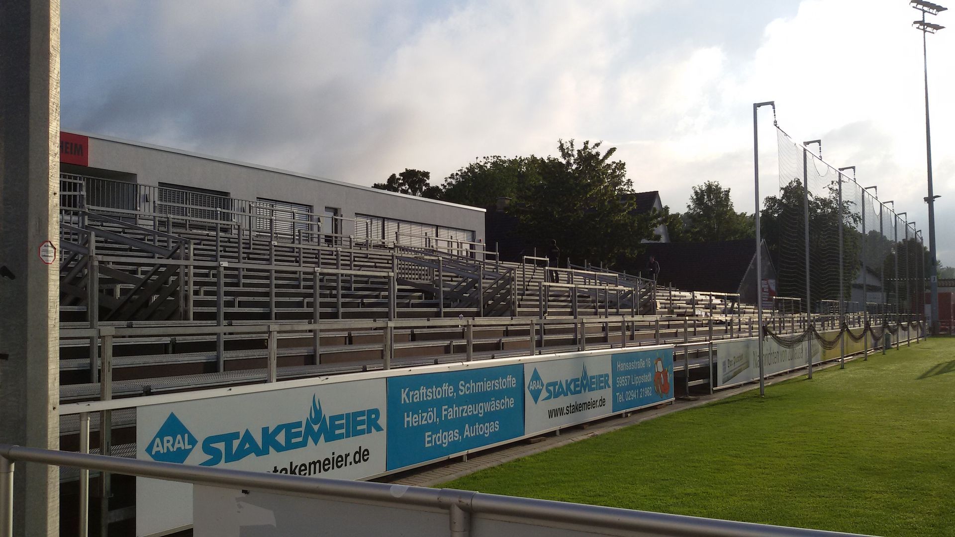 Leere Stadionsitze mit Werbetafeln und Netzen auf einem Rasenfeld, bewölkter Himmel.